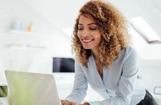 [Featured Image] A digital transformation specialist stands at her desk and uses her laptop to brainstorm ideas for reimagining a product for her organization. 
