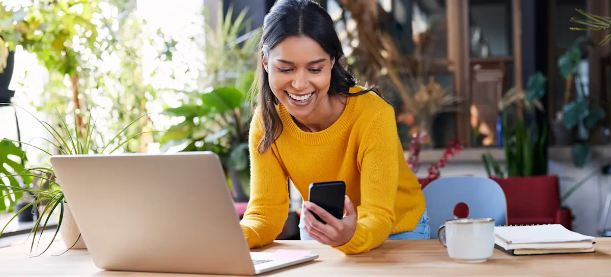 [Featured Image] A smiling person stands at a standing desk in their sunny home office using Google Veo to create a video from their laptop and checking it on their mobile phone.
