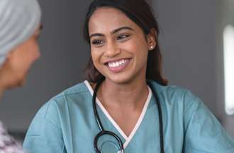 [Featured Image] An oncology nurse wearing light blue scrubs and a stethoscope around her neck speaks with her patient, who is wearing a black and white sweater and a head covering. 