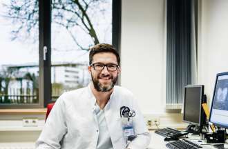 [Featured Image]:  A male, with short brown hair, a beard, and wearing glasses. He is wearing white pants, a white shirt, and a white jacket. He is sitting at his desk in front of his desktop and a larger screen on the wall. 
