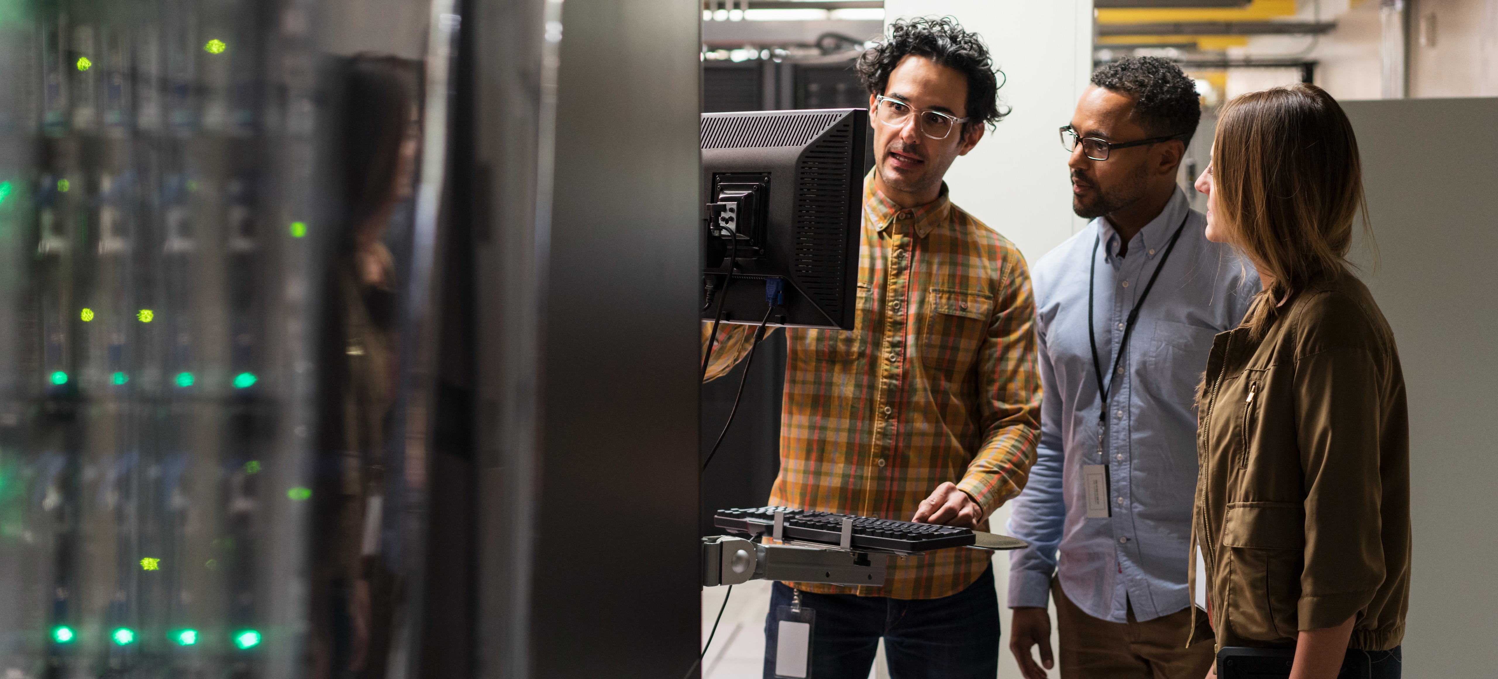 [Featured Image] A network administrator answers, "What does the term attenuation mean in data communication?" as they talk to two coworkers gathered around a desktop computer station in a networking room.