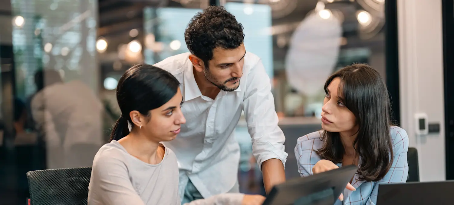 [Featured Image] Three colleagues sit in front of their laptops in an office setting and discuss cybersecurity best practices.

