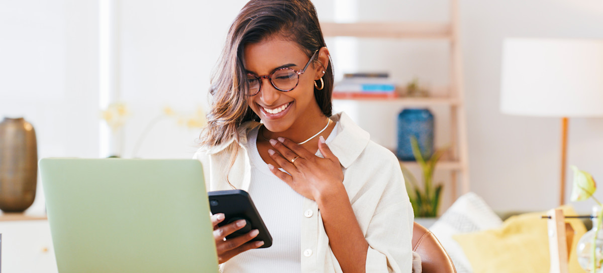 [Featured Image] A college student sitting in front of their laptop looks at their phone, receiving good news after choosing between early action and early decision.
