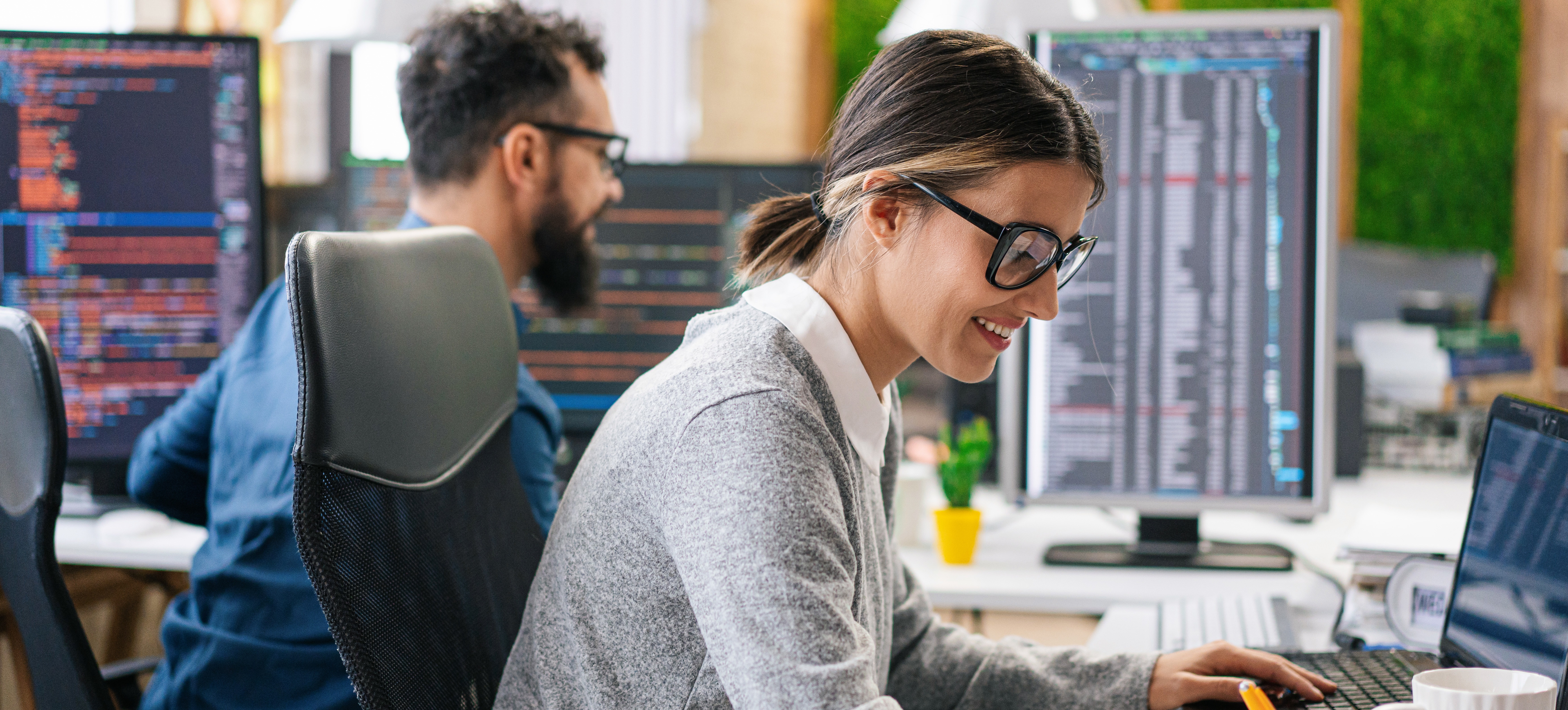 [FEATURED IMAGE] Two full-stack web developers share a desk and look at their desktop computer screens. 
