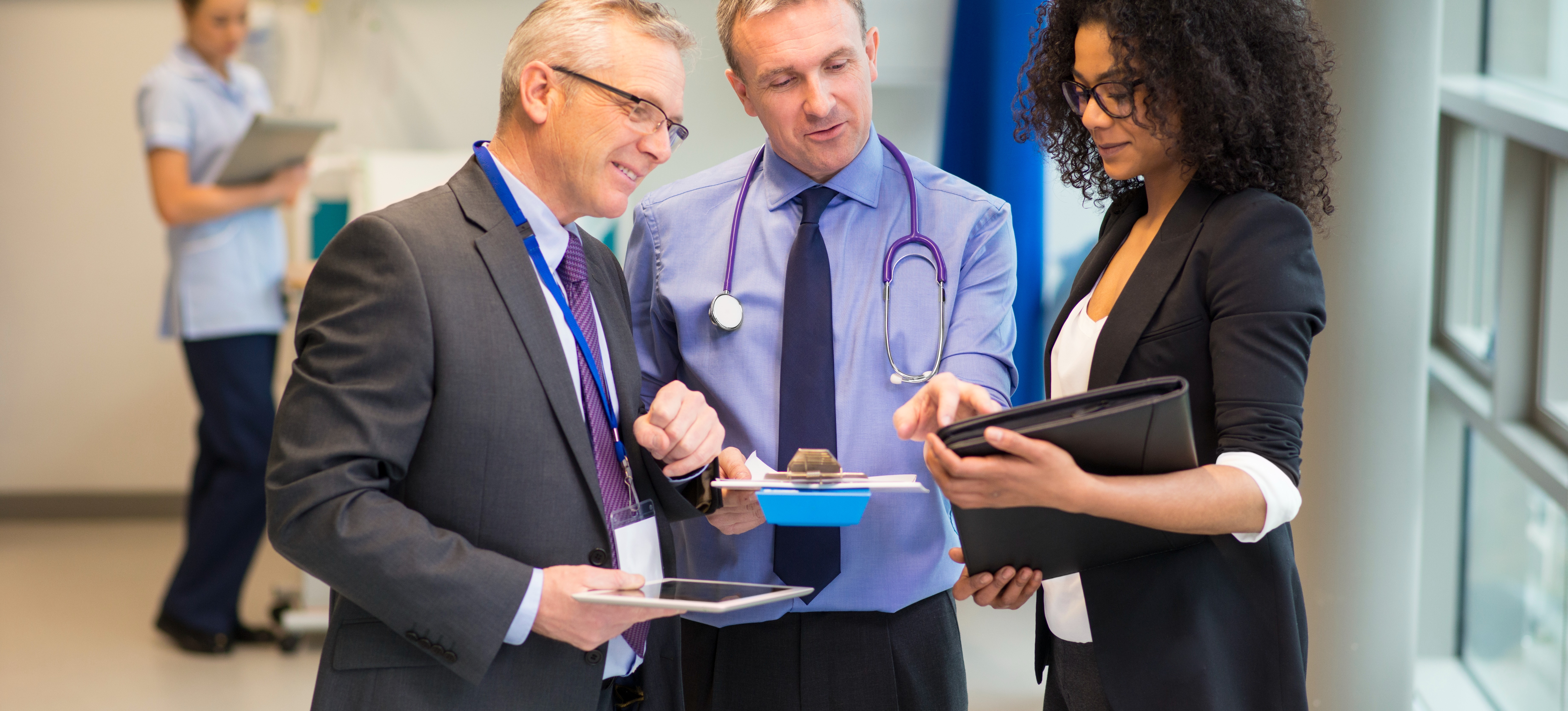 [Featured Image] Two hospital administrators, who have earned a healthcare MBA, stand in a hospital corridor with a doctor discussing information written on a notebook that one of the administrators is holding.