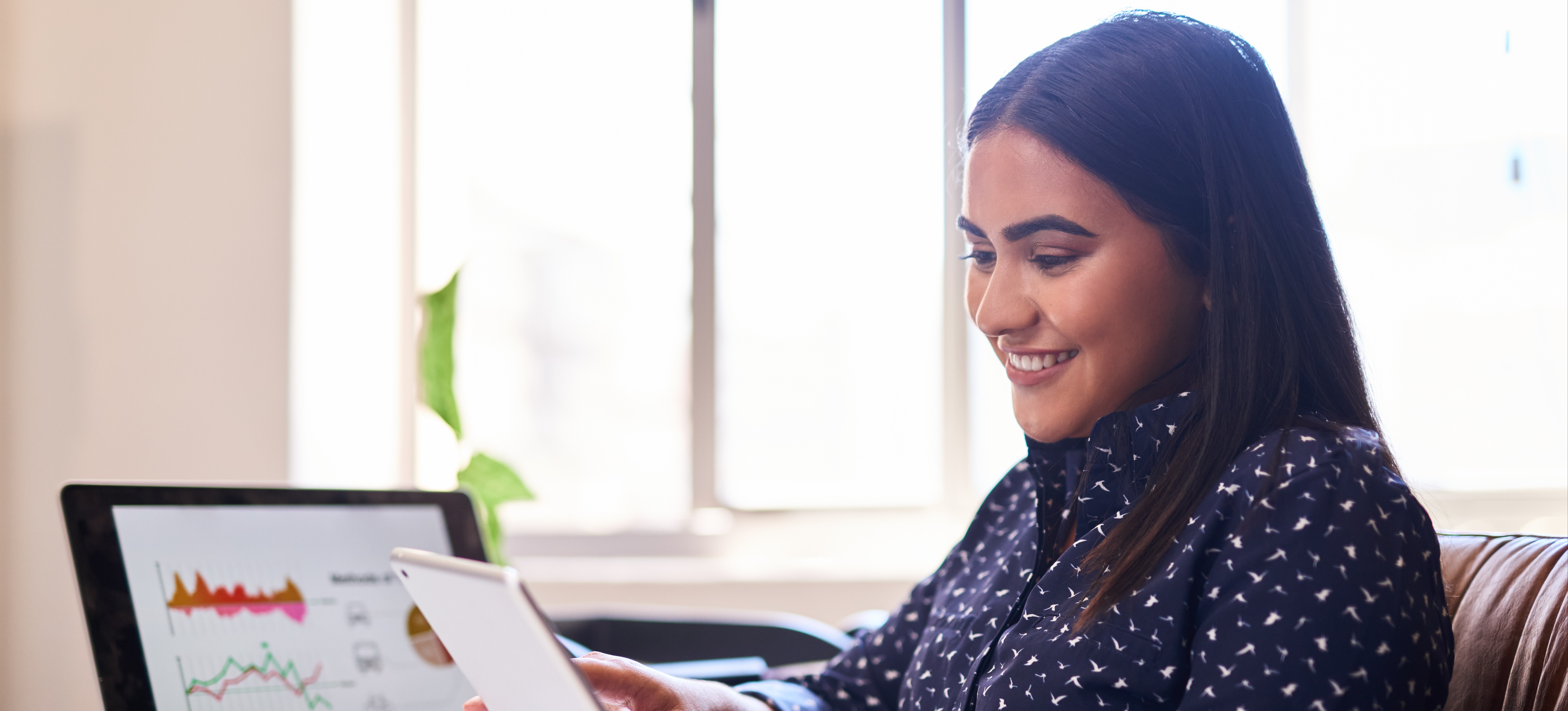 [Featured Image] A smiling data scientist assesses data on a tablet while referencing charts on a laptop in their office.
