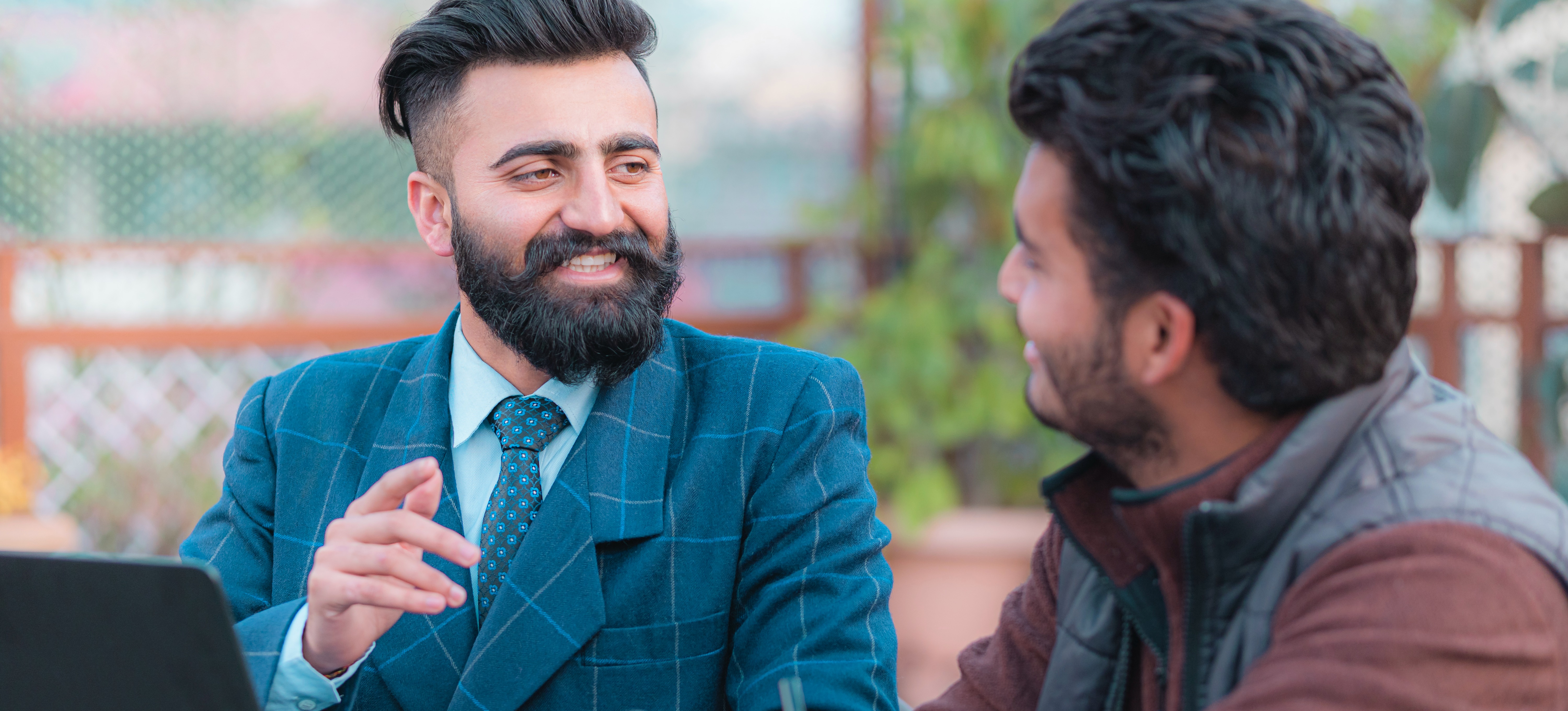 [Featured Image] A management consultant dressed in a blue suit and tie discusses business with a client while they are sitting in an outdoor venue in front of a laptop.
