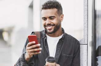 [Featured Image] A man stands outside a building holding a to-go coffee and listening to a podcast on his phone.