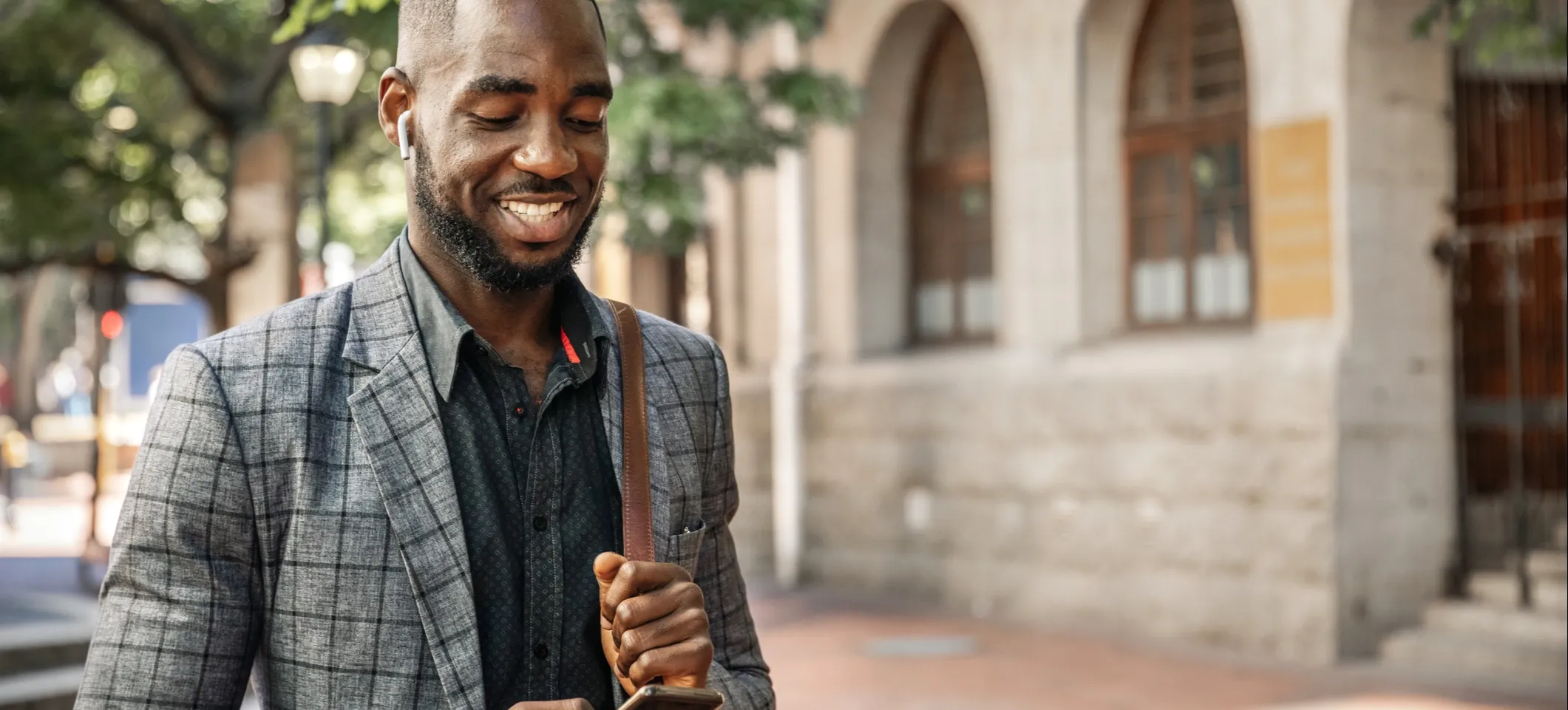 [Featured image] A master's degree student in a grey and black striped jacket and button-up shirt looks at their cellphone while walking across a college campus.
