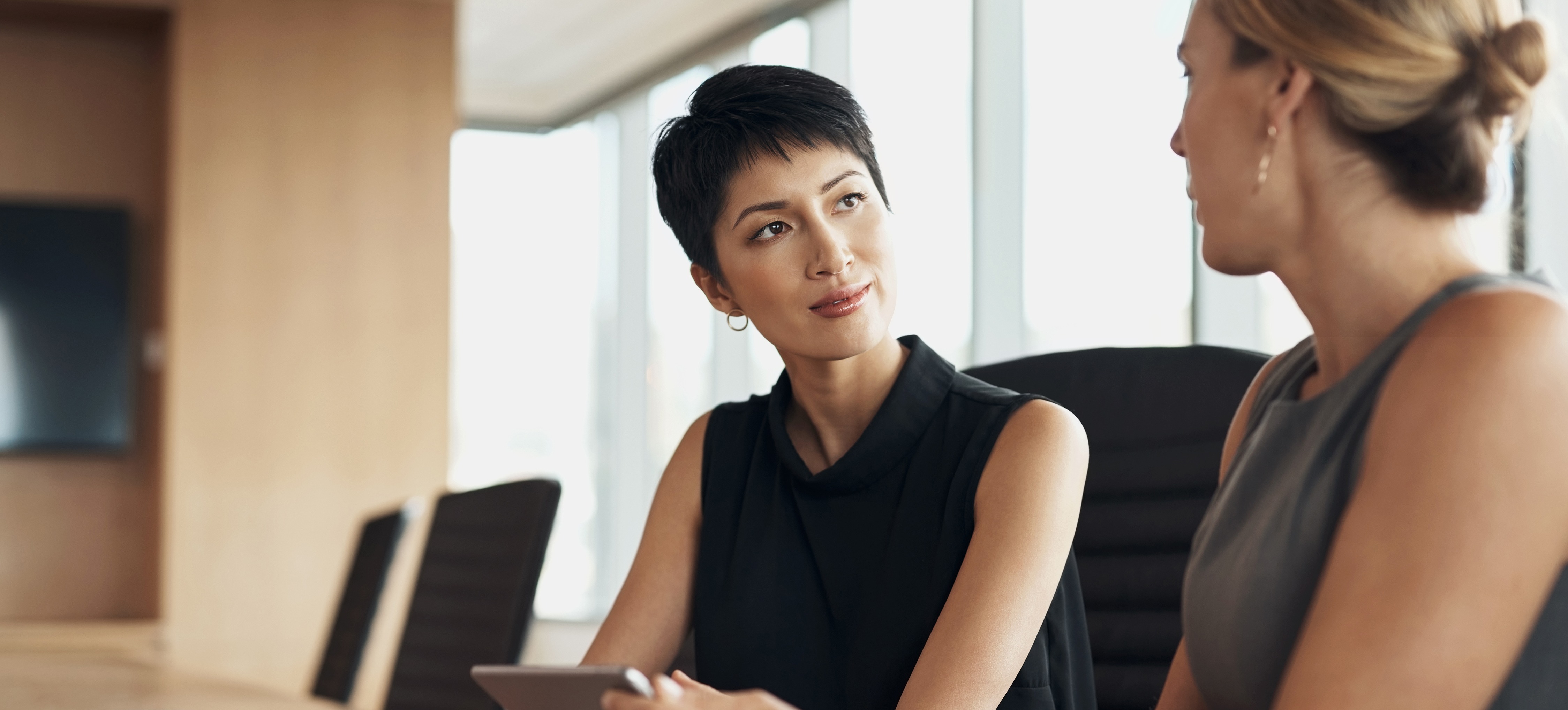 [Featured Image] A business person who embodies what is a CEO sits at a long boardroom table and listens to their colleague speak.
  
