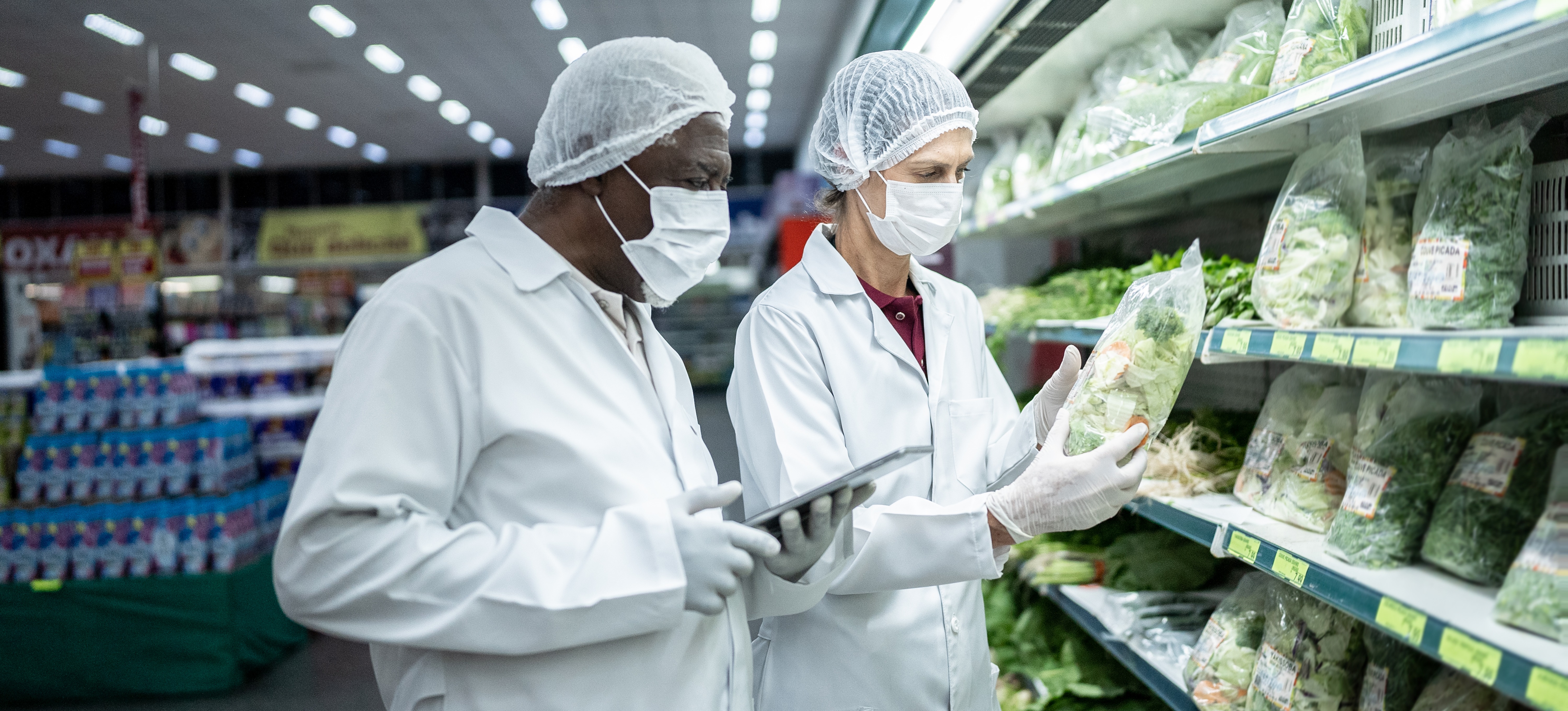 [Featured Image] Two health inspectors dressed in white lab coats, gloves, face masks, and hair nets stand in the produce section of a grocery store and inspect sealed bags of lettuce.

