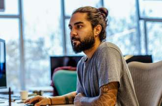 [Featured image] A tattooed male computer technician works at a dual-screen workstation in an office with windows behind him. 