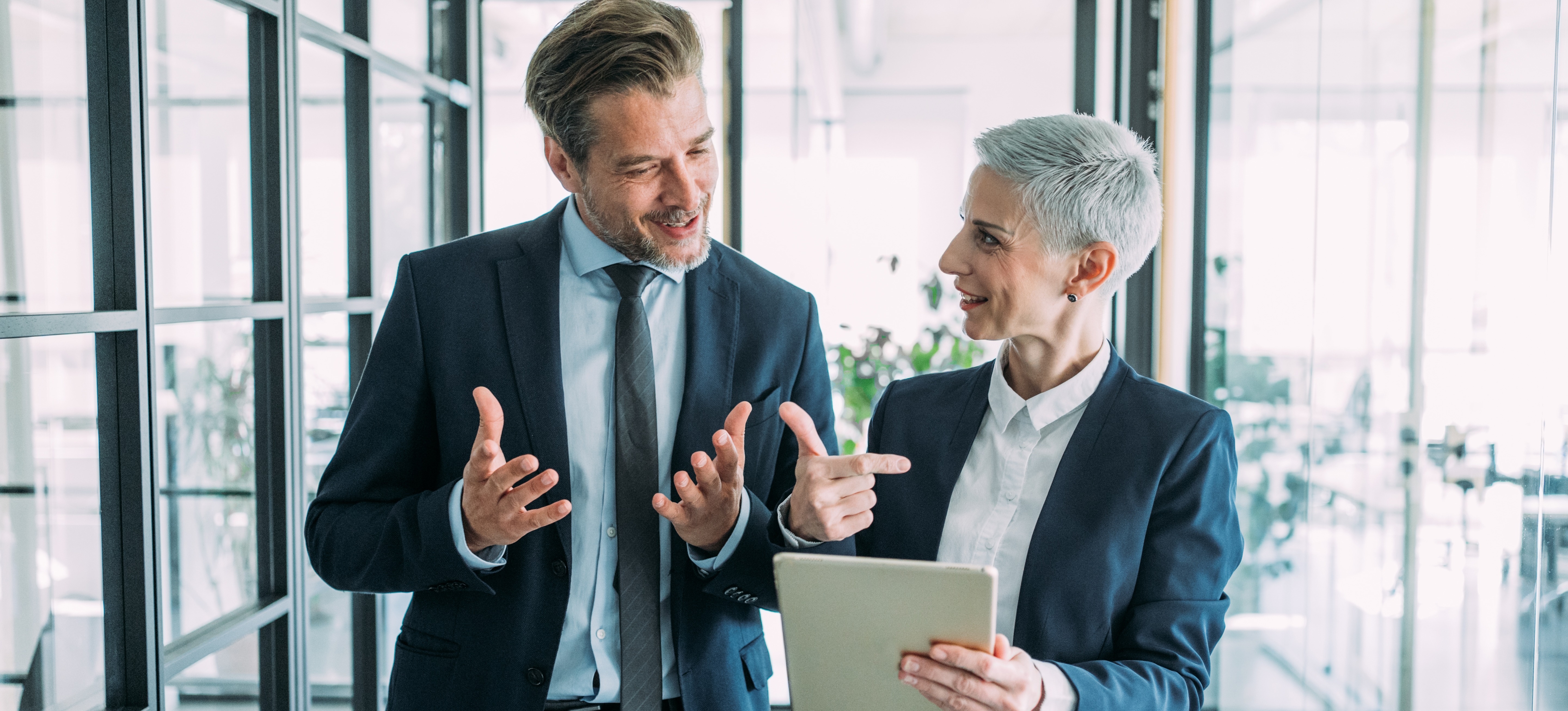 [Featured Image] A technical consultant holds a tablet while talking with a colleague as they walk through an office.
