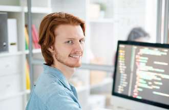 [Featured Image]: A male, with red hair, beard, mustache,  and wearing a blue shirt, is sitting in front of his desktop, in his office.