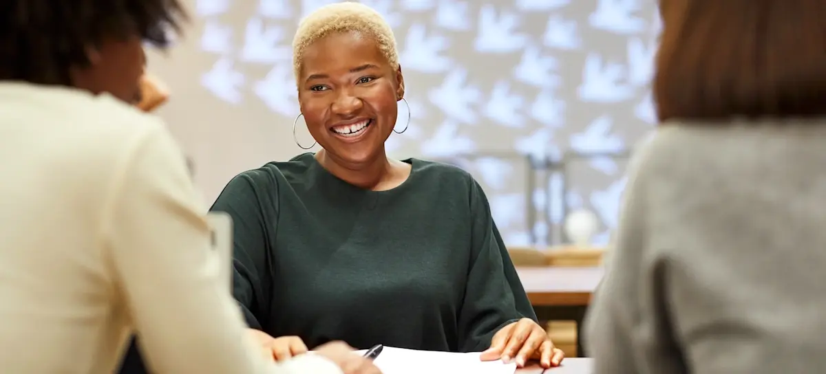 [Featured Image] A smiling worker with short hair and earrings sits at a table answering UX designer interview questions.