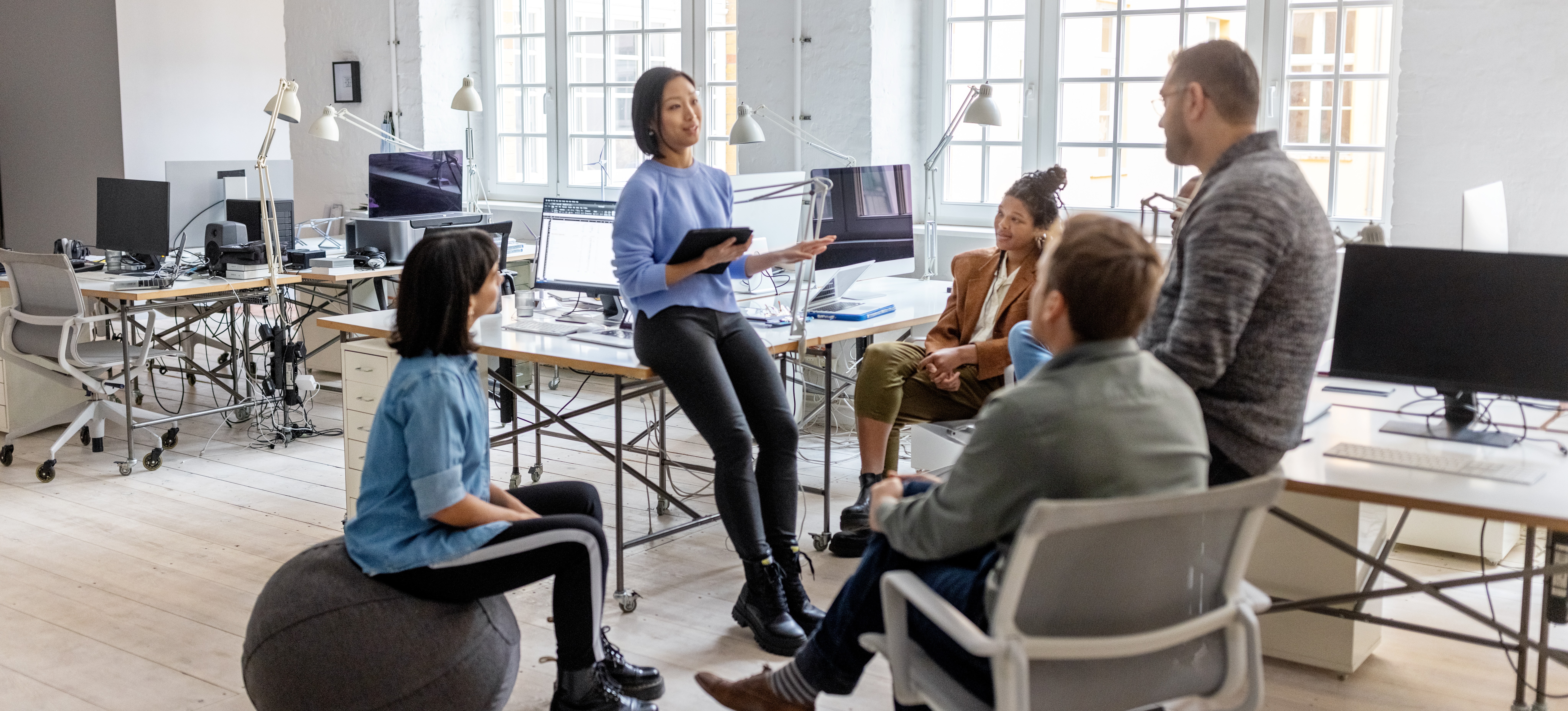 [Featured Image] A project manager, who has earned her project management certification, speaks with her team in an office setting.
