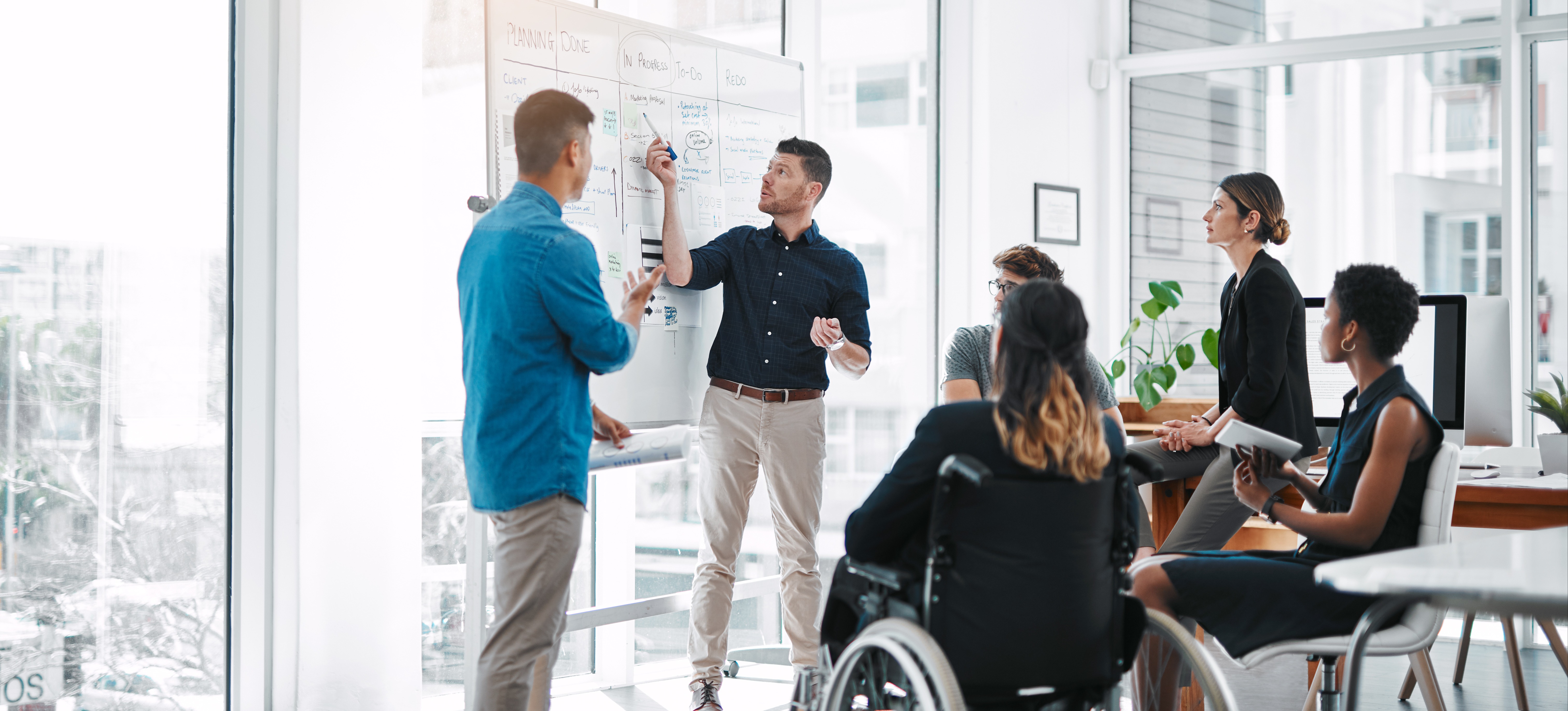 [Featured Image] A marketing team discusses their funnel marketing strategies, with one person pointing to a whiteboard while their colleagues watch.
