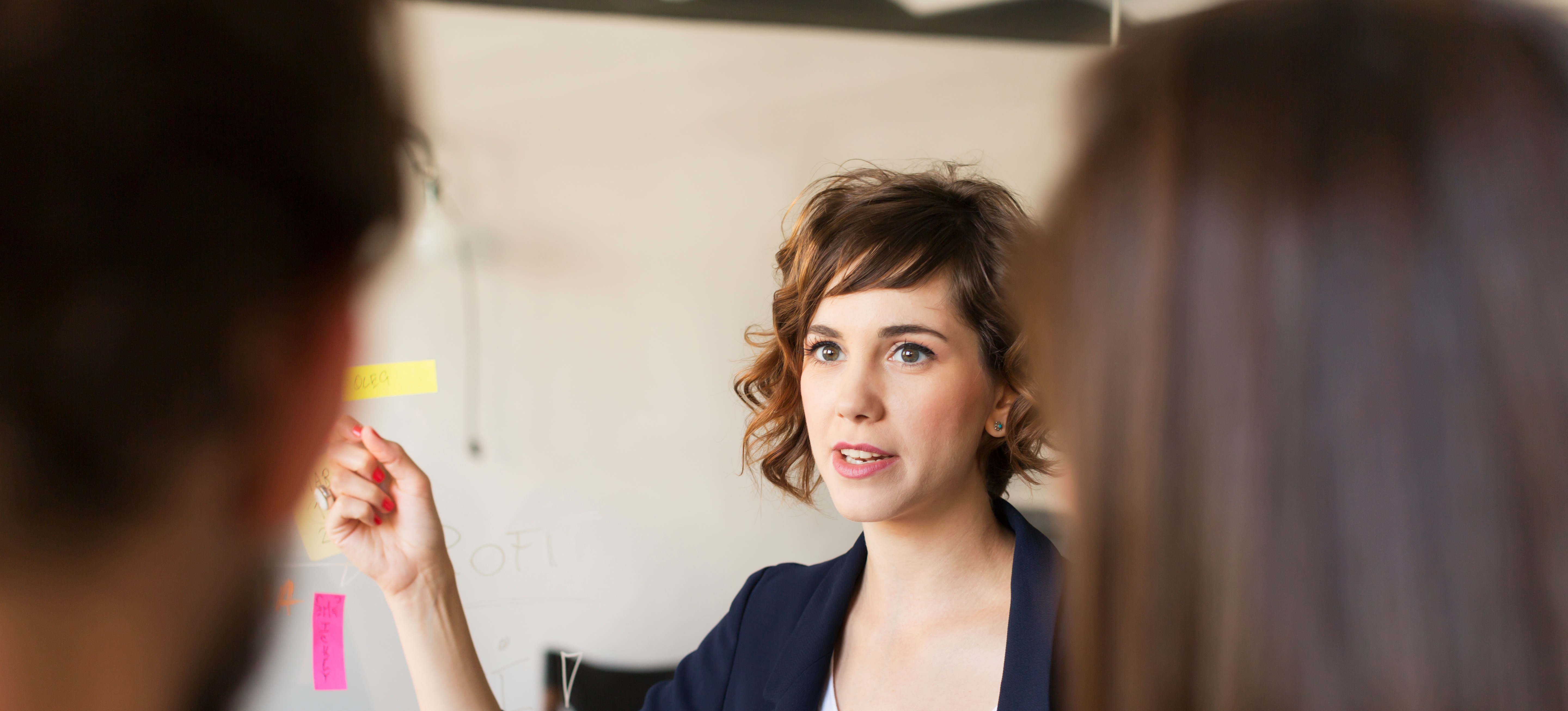 [Featured image] A professional, wearing a dark jacket and white top, is standing in front of and pointing to a whiteboard, as they conducts a meeting with the product development team.