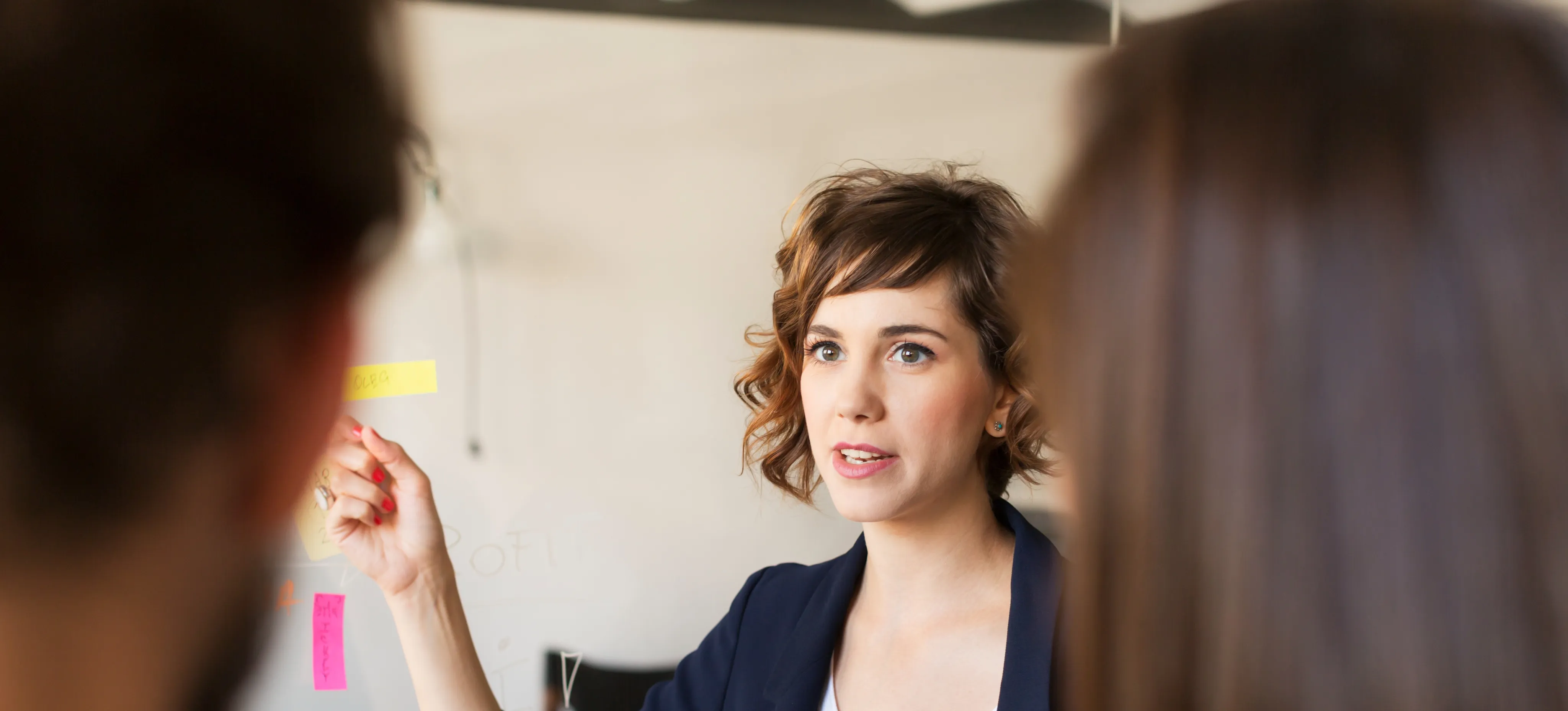 [Featured Image] A professional, wearing a dark jacket and white top, is standing in front of and pointing to a whiteboard, as they conduct a meeting with the product development team.