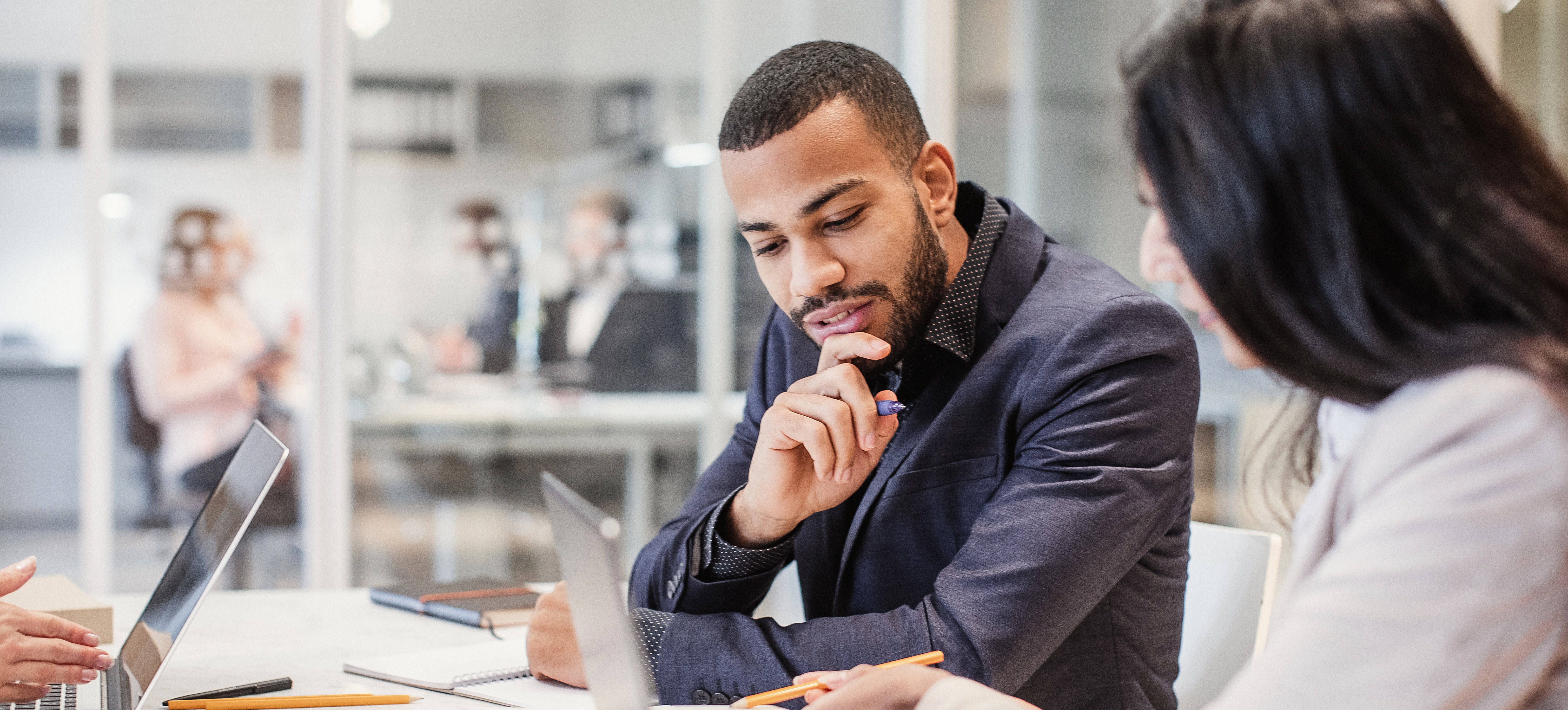 [Featured Image] A man who followed an HR career path sits in a meeting next to a colleague and looks over at something in her notebook.
