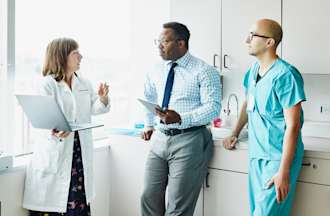 [Featured Image] Female doctor leading medical team discussion in a hospital exam room.