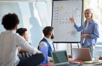 [Featured image] A woman in a blue shirt shows a marketing plan on a whiteboard to a group.