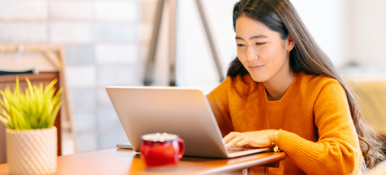 [Featured image] A cloud engineer wearing an orange sweater works at her laptop with a hot beverage in an office.