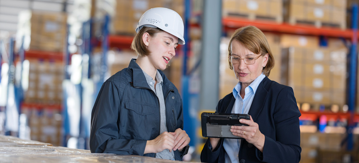 [Featured Image] A supply chain analyst holding a device records and discusses information with a warehouse worker while they stand in the warehouse.
