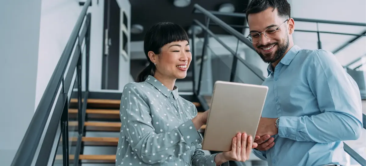 [Featured Image] A smiling supervisor shows an employee their performance management goals on a device as they stand in an office.