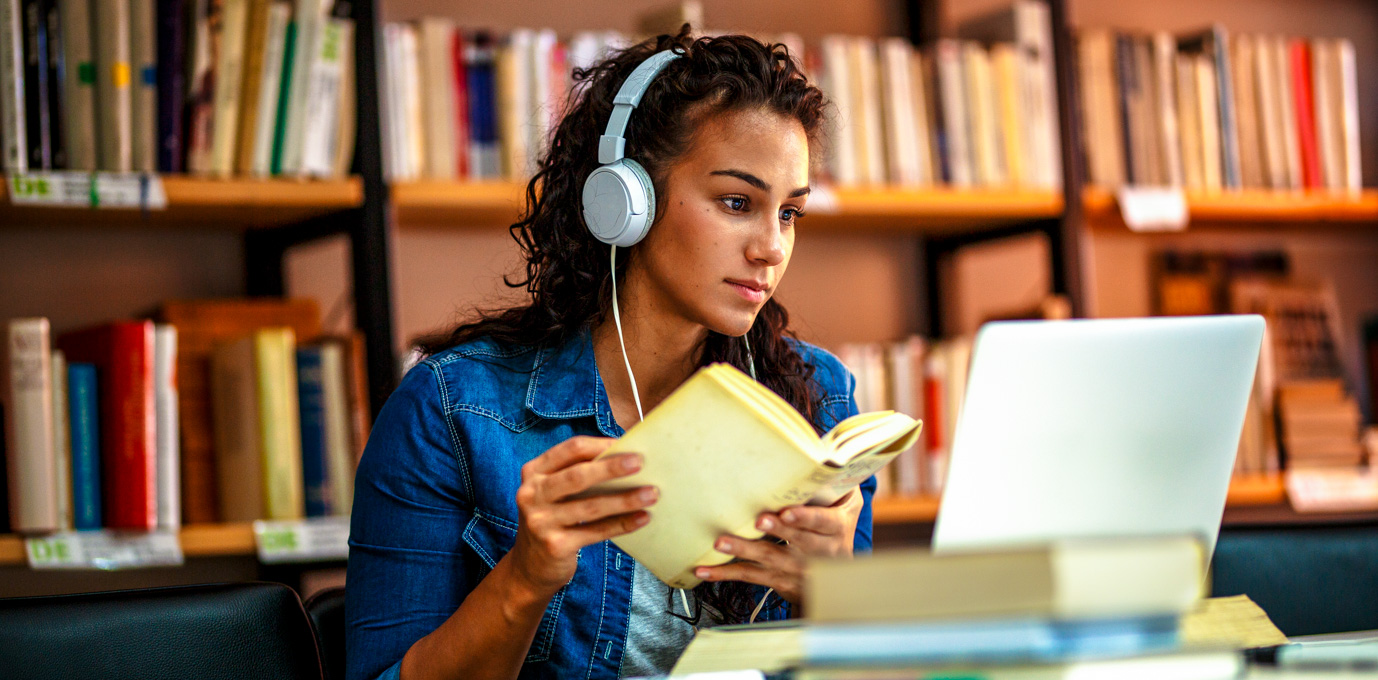 [Featured Image] A college student wears headphones and sits at a library table with a book in their hands and a laptop in front of them.