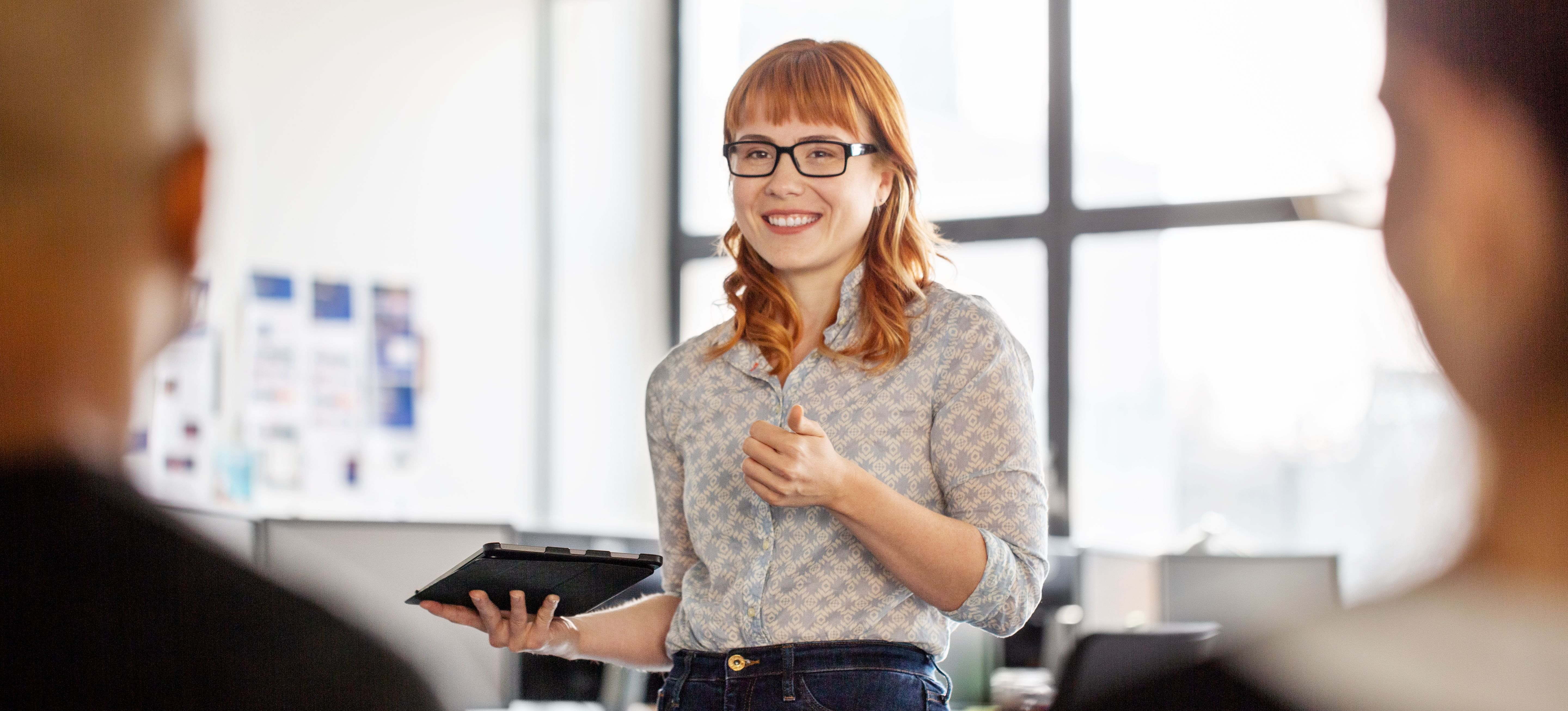 [Featured Image] A smiling project manager is in business casual clothing and is facing two coworkers while holding a tablet. 