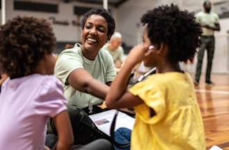 [Featured image] A community health worker sits in a gymnasium working with two young children.