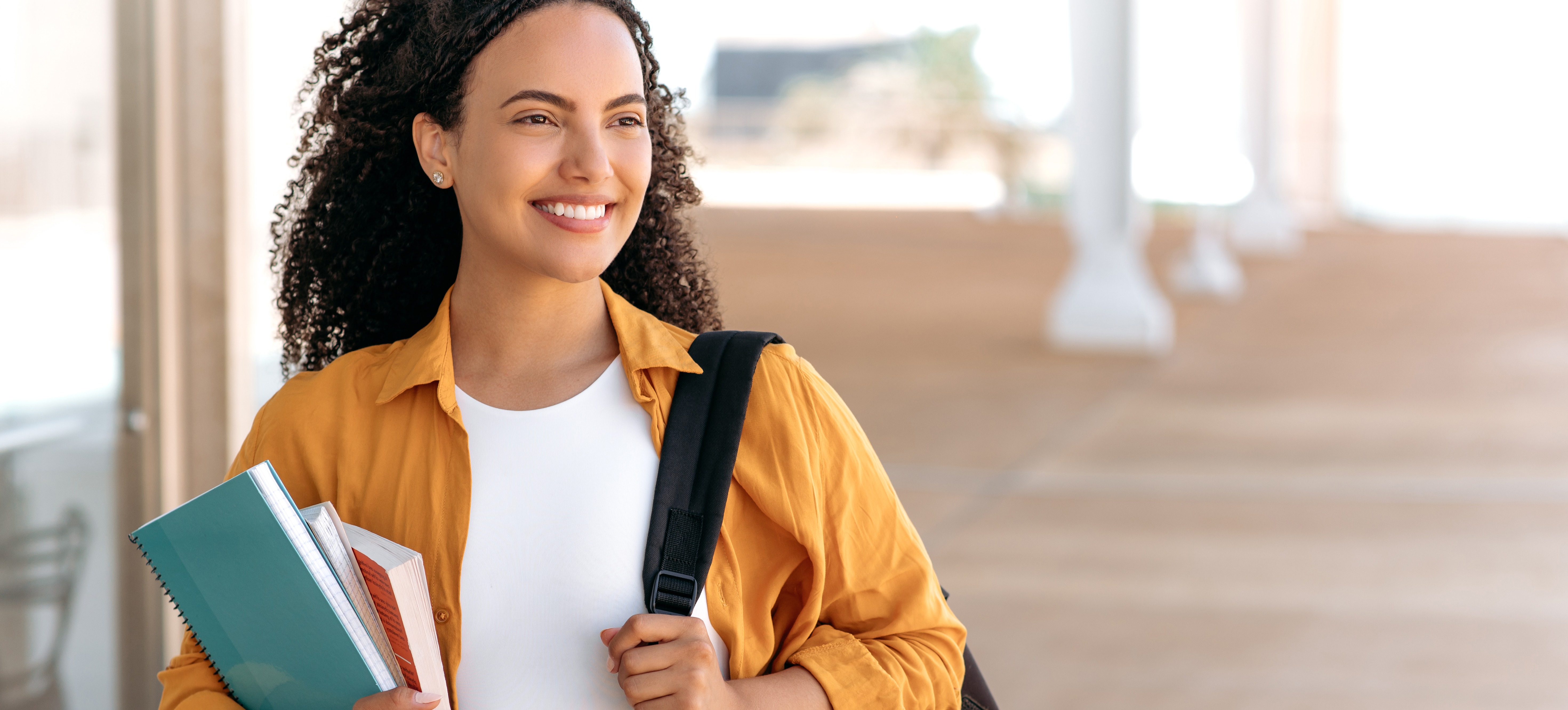 [Featured Image] A curly-haired student with a backpack holds books and notebooks in their hand and walks on a college campus.