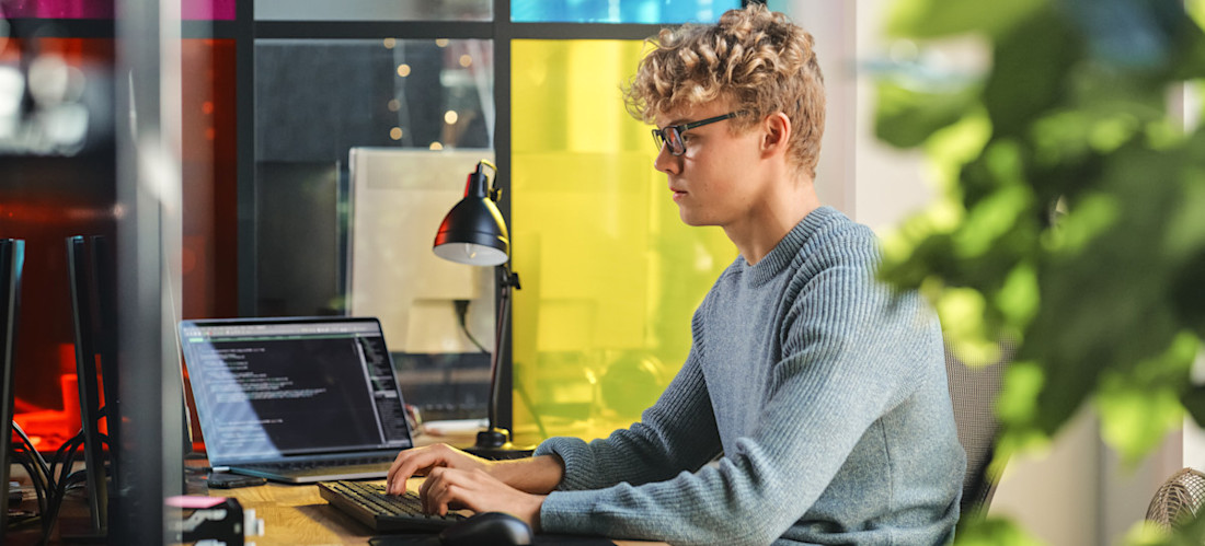 [Featured Image] A young programmer uses the Julia programming language as they work on code at two computers in a colorful, casual office setting.
