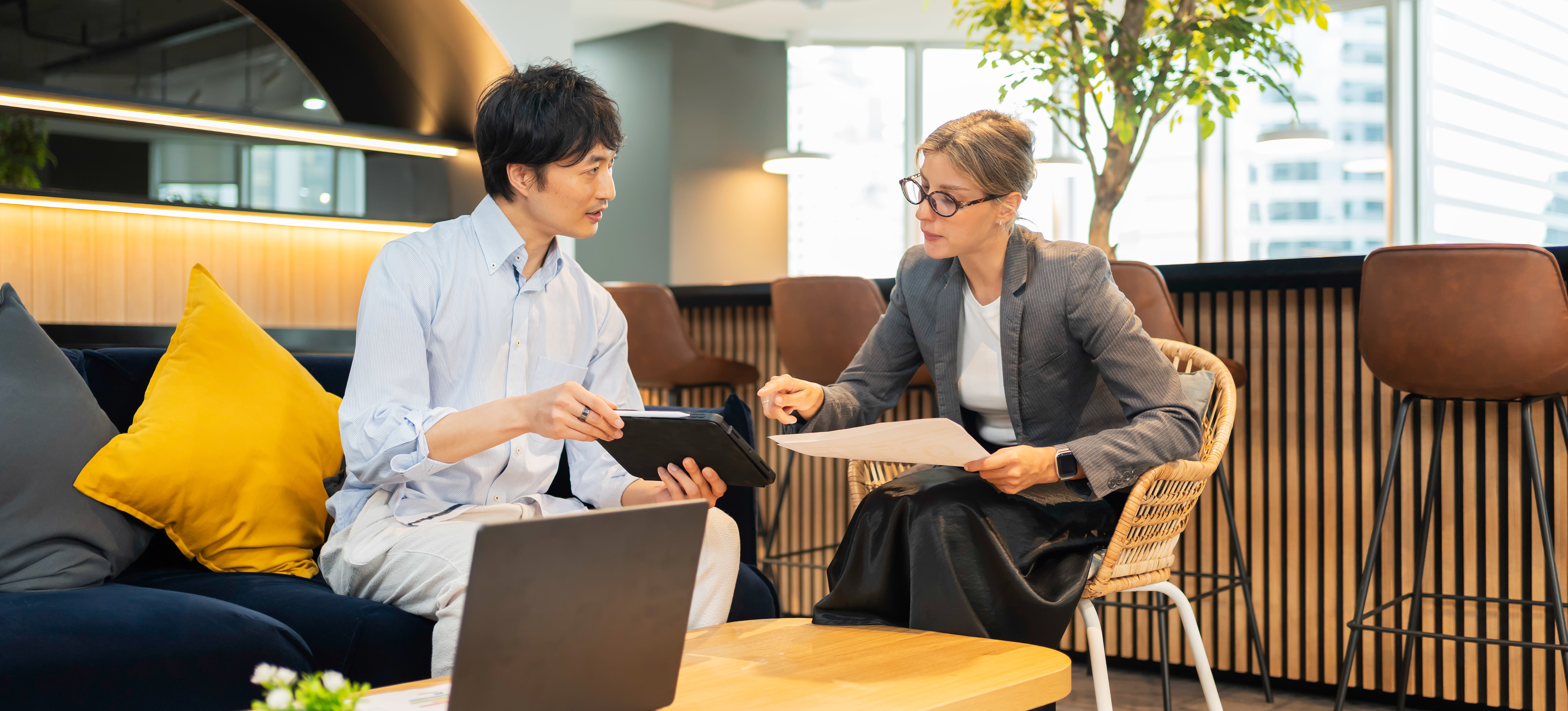 [Featured Image] Two colleagues discuss how using AI for scheduling streamlines operations and boosts productivity in their workplace while reviewing tool options on a tablet in a break area.
