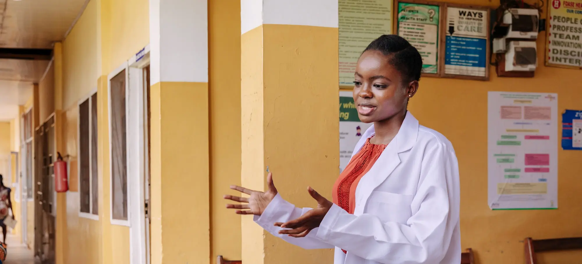 [Featured Image]:  A health services manager, wearing a white lab coat and orange top, standing outside an office, preparing to meet with hospital employees.