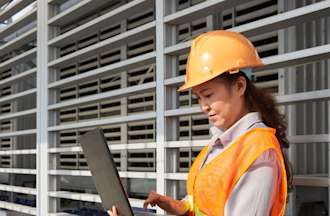 [Featured Image] An electrical engineer in an orange helmet and safety vest reviews charts and information on the job site. 