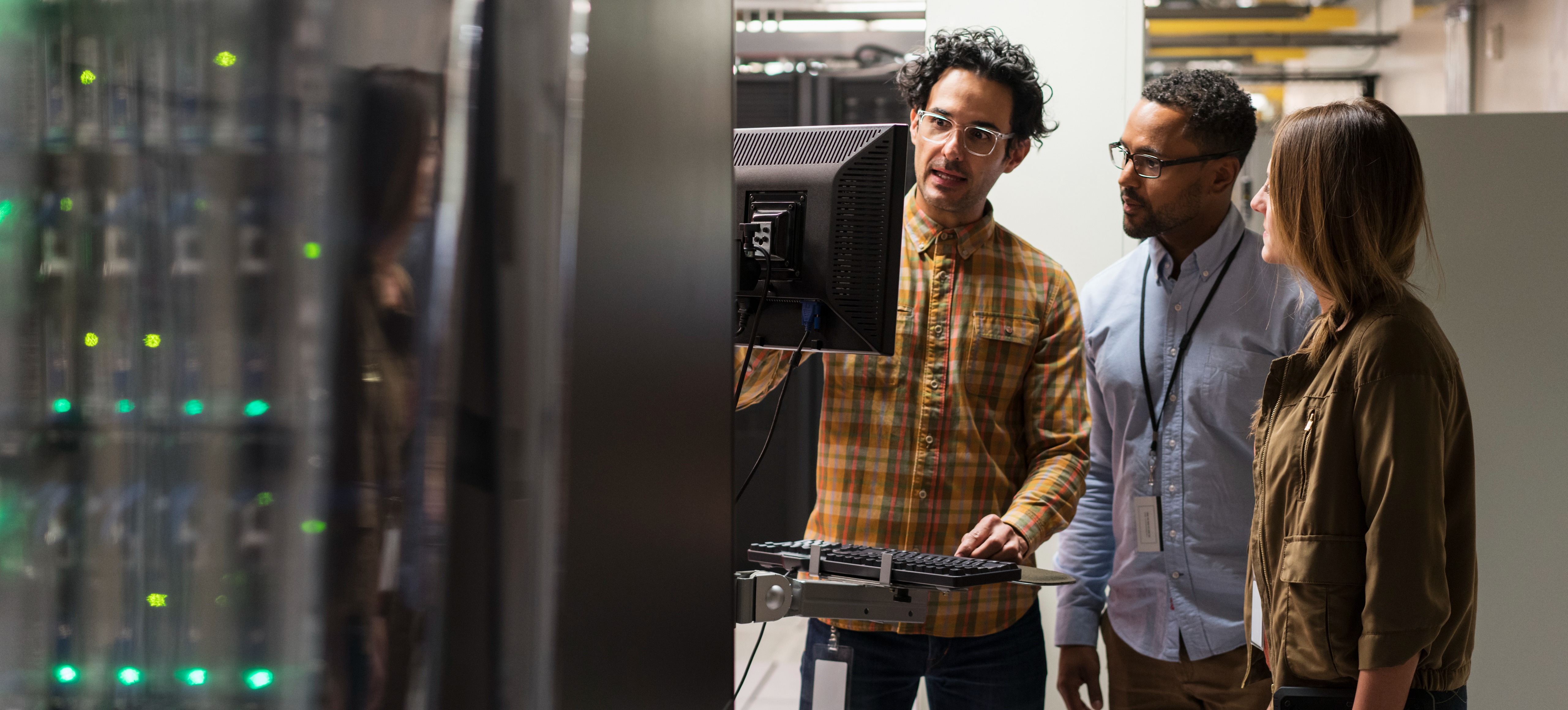 [Featured image] Three colleagues knowledgeable of IT terms stand together in a server room and troubleshoot an issue on a computer monitor. 