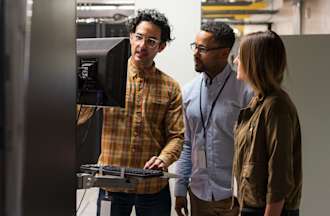 [Featured image] Three colleagues stand together in a server room and troubleshoot an issue on a computer monitor. 