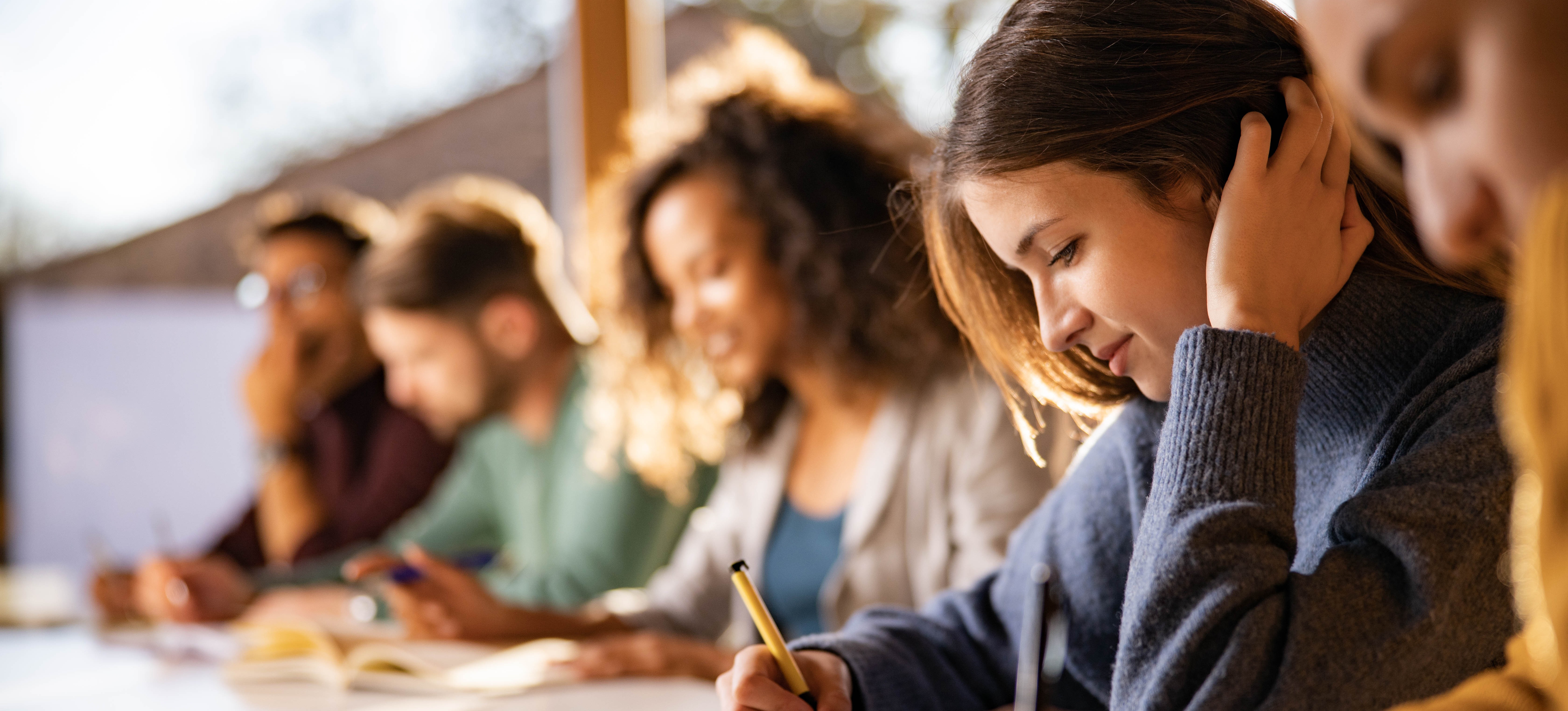 [Featured Image] After paying their college tuition, a row of students sit working in class, writing in notebooks.
