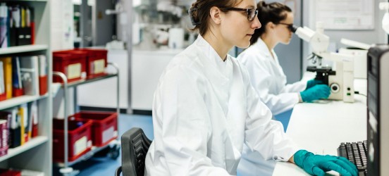 [Featured image] Two medical lab technicians in white coats and gloves work in a medical lab. One is typing on a computer, and the other is looking at a specimen under a microscope.