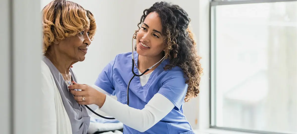 [Featured Image] A smiling nurse practitioner listens to a patient's heartbeat with a stethoscope as they both sit on a hospital bed.