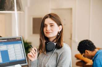 [Featured image] A design manager with long brown hair and wearing a gray sweatshirt, is standing in front of her desktop in her office. She is wearing a headphone around her neck.