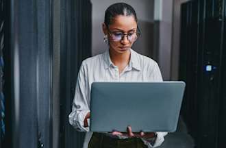 [Featured Image] A woman holds a laptop computer in a server room. 