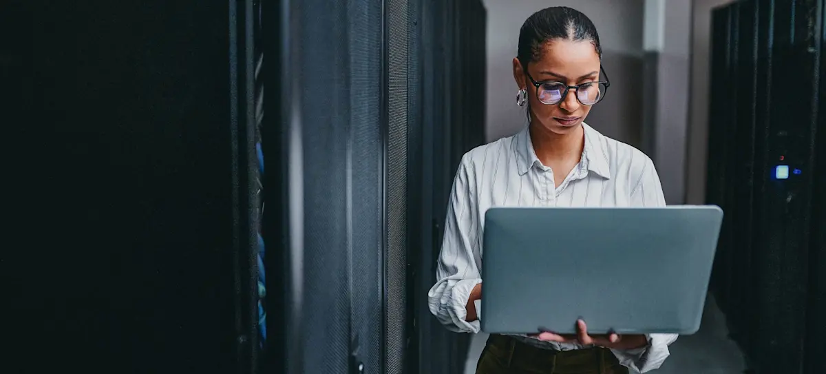 [Featured Image] A worker holds a laptop computer in a server room. 