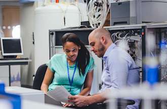 [Featured Image] Two power electronics engineers examine documents in front of electrical equipment.