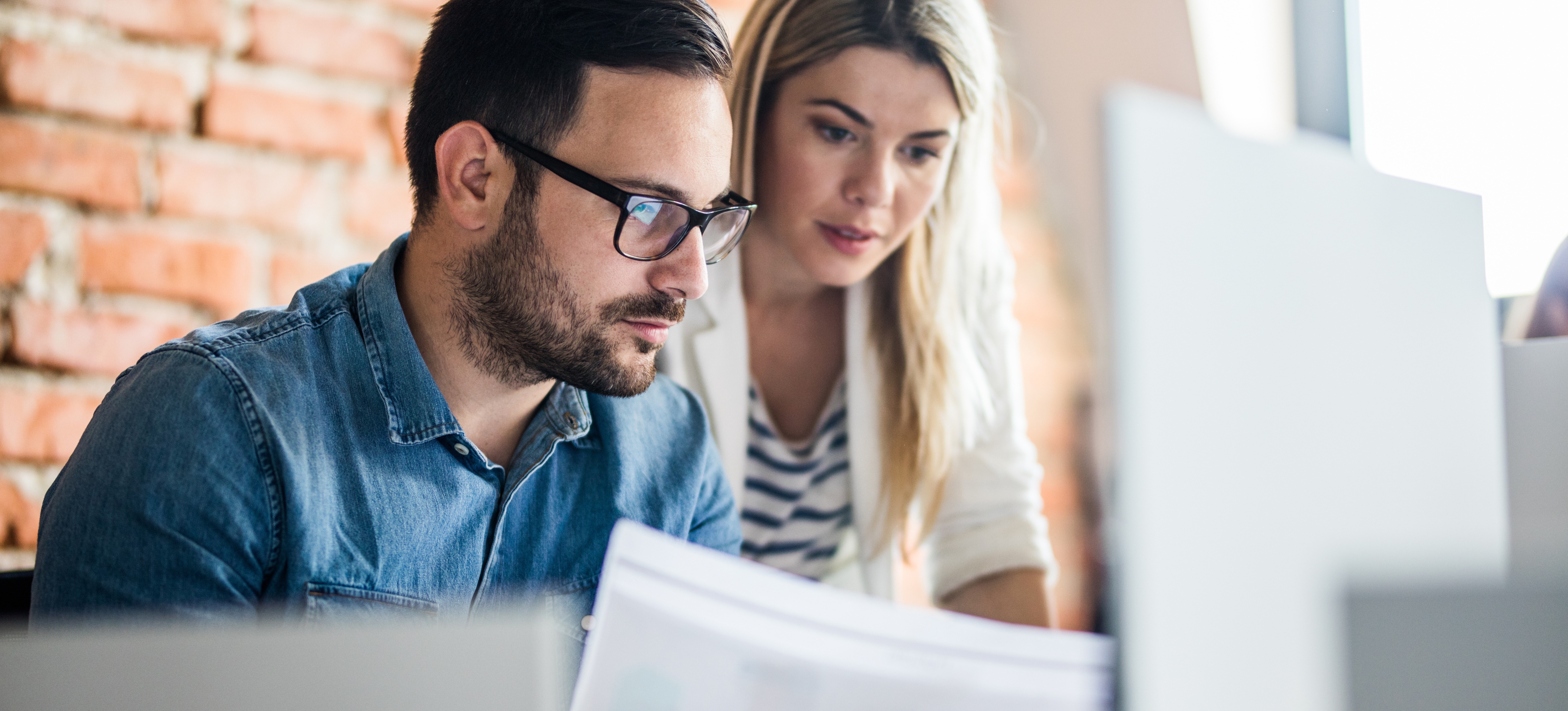 [Hauptbild] Zwei Geschäftskollegen, ein Mann und eine Frau, analysieren die Daten auf einem Blatt Papier und einem Computermonitor, die mit einem Business-Intelligence-Tool erfasst wurden.
