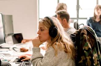 [Featured Image] An entry-level programmer examines code on a computer monitor beside a laptop.