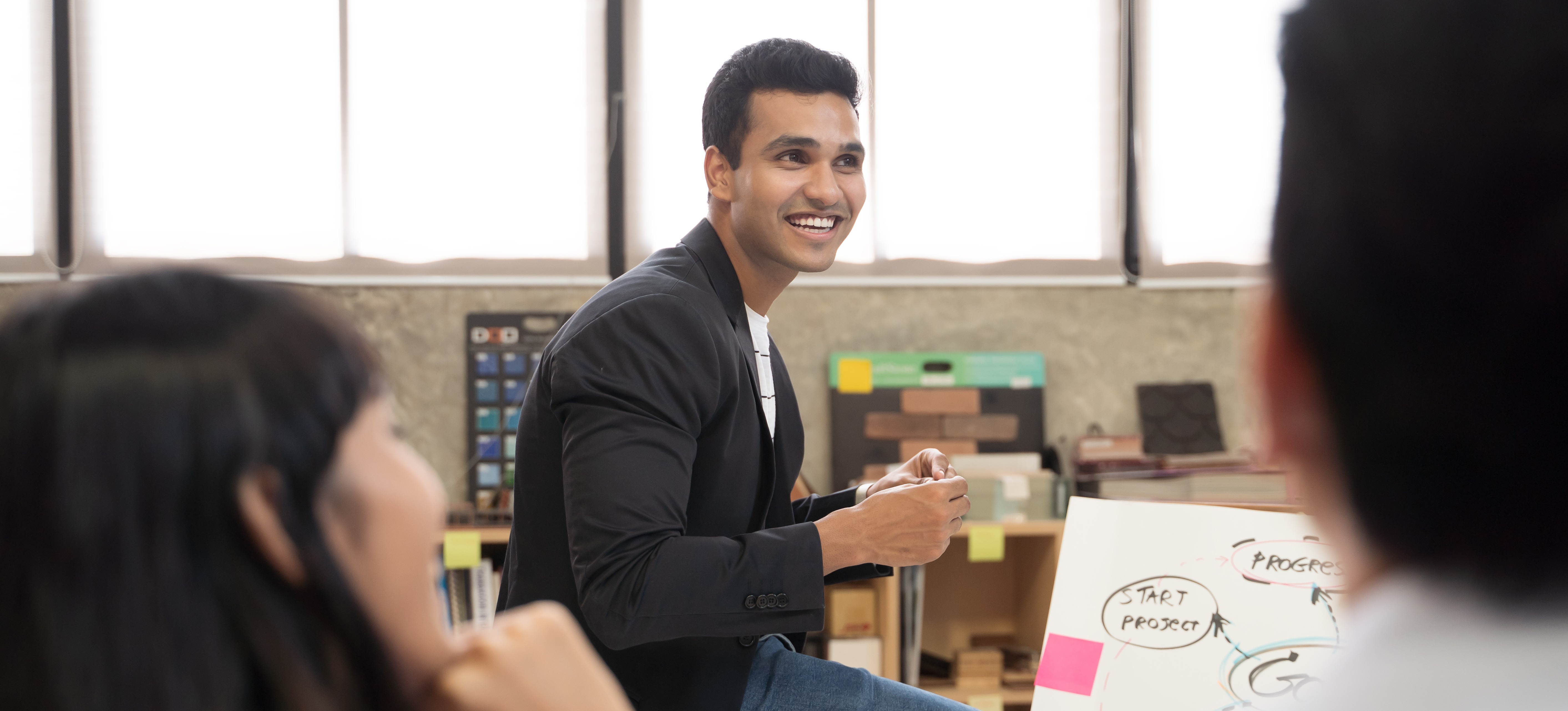 [Featured Image] A certified project manager sits on the edge of a worktable whilst discussing the benchmarks and progress of the team’s latest project. 

