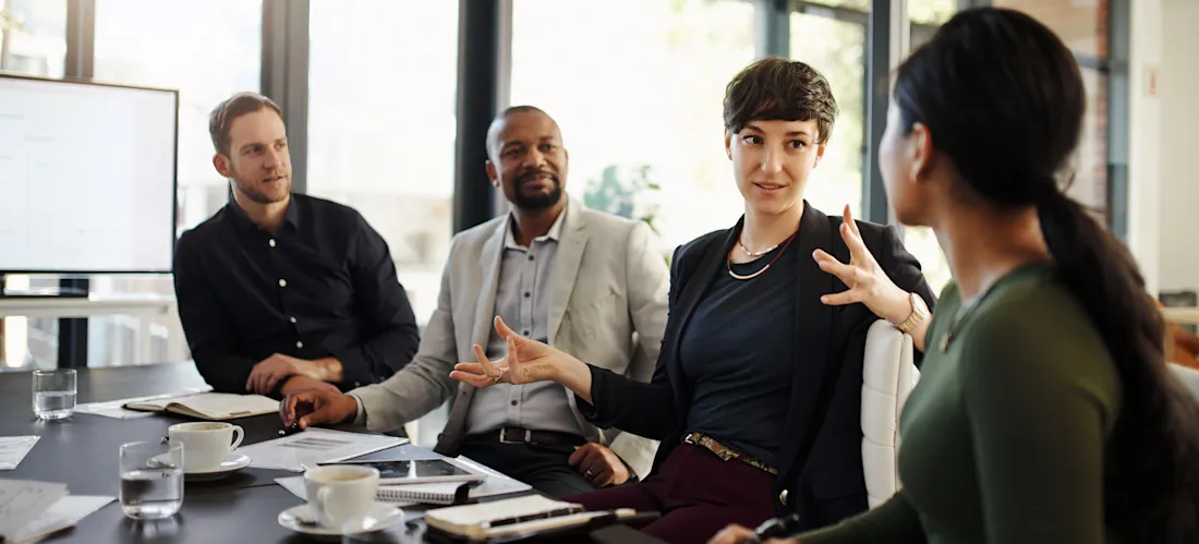 [Featured image] Four colleagues sit at a conference table discussing business marketing.